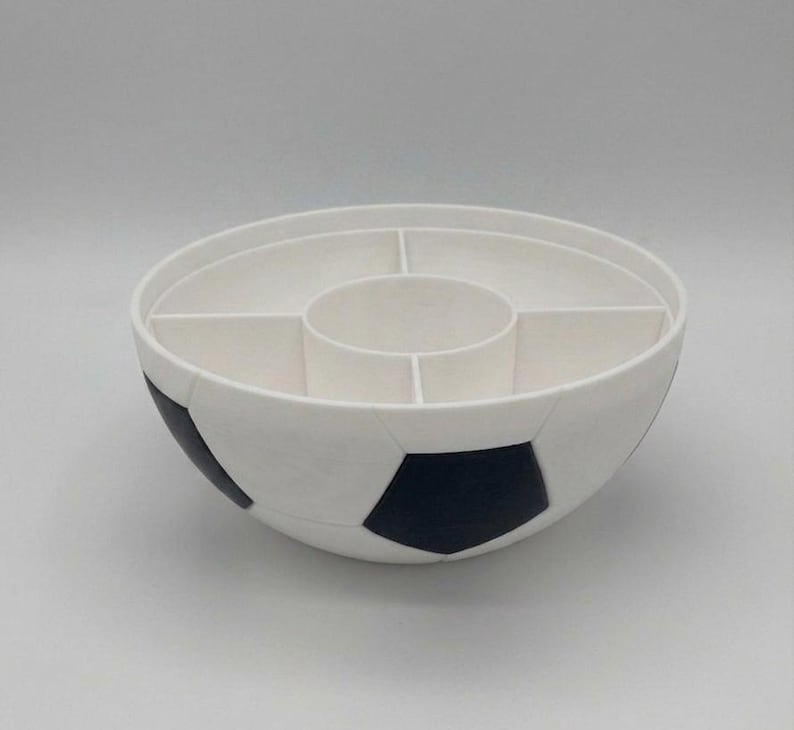 May include: A white and black soccer ball shaped serving bowl with a divided interior. The bowl features a central circular compartment surrounded by four outer sections, mimicking the classic pentagonal pattern of a soccer ball on the exterior.