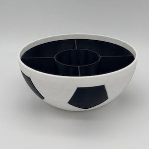 May include: A round bowl designed with a black and white soccer ball pattern on the exterior. The interior is black and features a central circular compartment surrounded by four additional curved sections, creating a divided storage container.
