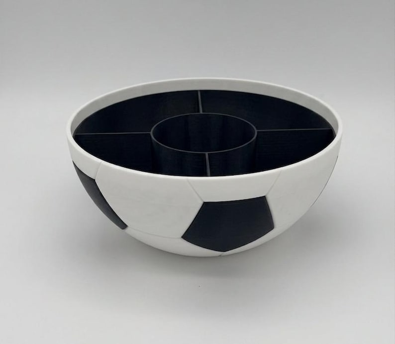 May include: A round bowl designed with a black and white soccer ball pattern on the exterior. The interior is black and features a central circular compartment surrounded by four additional curved sections, creating a divided storage container.
