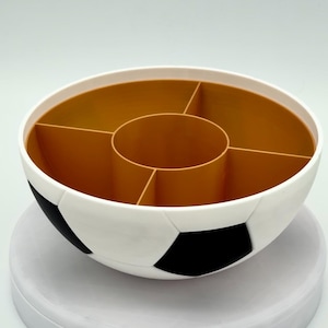 May include: A round, bowl-shaped organizer designed with a soccer ball exterior featuring a classic black and white pentagonal pattern. The interior is a solid tan color and is divided into five distinct sections, including a central circular compartment.