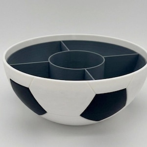 May include: A round bowl with a white and black soccer ball pattern on the exterior. The interior is dark gray and features a central circular compartment surrounded by four separate wedge-shaped sections for organized storage and serving.