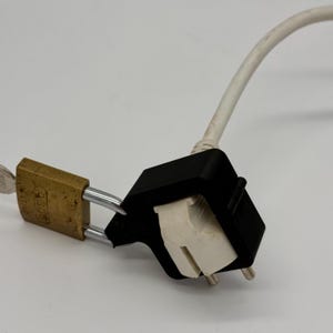 Könnte beinhalten: Ein weißes Stromkabel mit einem schwarzen Stecker aus Kunststoff ist mit einem Messing-Vorhängeschloss gesichert. Das Vorhängeschloss trägt die Aufschrift "Master".