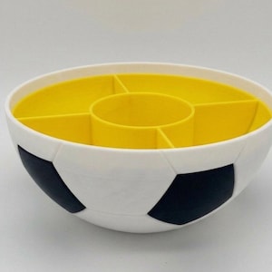 May include: A round, white bowl with a black soccer ball pattern on the exterior. The interior is bright yellow and features a central circular compartment surrounded by four separate wedge-shaped sections for organized storage or serving.