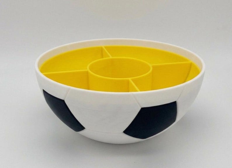 May include: A round, white bowl with a black soccer ball pattern on the exterior. The interior is bright yellow and features a central circular compartment surrounded by four separate wedge-shaped sections for organized storage or serving.