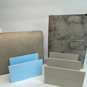 Könnte beinhalten: Ein hellblaues rechteckiges Namensschild mit dem geprägten Namen Alexandra, ein beiges rechteckiges Namensschild mit dem geprägten Namen Alexandra, eine einfarbige beige Clutch und ein graues Notizbuch mit Marmormuster und Druckknopf.