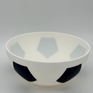 May include: A white bowl featuring a black and gray pentagonal pattern on its exterior and interior, designed to resemble the classic aesthetic of a soccer ball. The bowl has a smooth, rounded shape with a wide opening and a solid base.