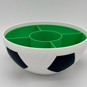 May include: A round serving bowl designed with a soccer ball pattern on the exterior, featuring black and white pentagonal shapes. The interior is bright green and divided into five separate compartments, including a central circular section.