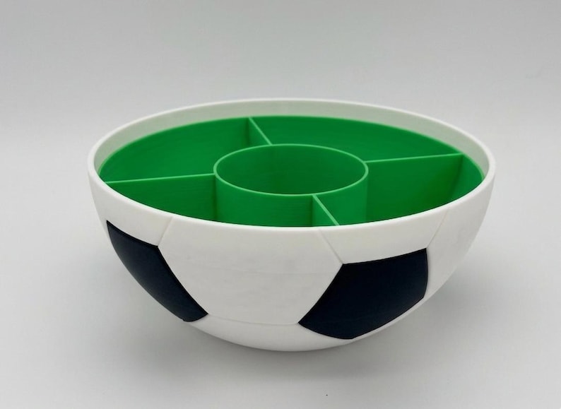 May include: A round serving bowl designed with a soccer ball pattern on the exterior, featuring black and white pentagonal shapes. The interior is bright green and divided into five separate compartments, including a central circular section.