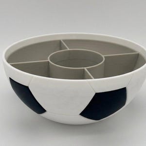 May include: A round bowl designed to resemble a soccer ball with a white and black exterior pattern. The interior is light gray and features a central circular compartment surrounded by four additional divided sections for organized storage.