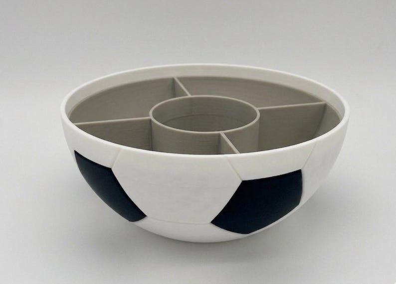 May include: A round bowl designed to resemble a soccer ball with a white and black exterior pattern. The interior is light gray and features a central circular compartment surrounded by four additional divided sections for organized storage.