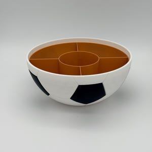 May include: A round, bowl-shaped snack organizer designed with a soccer ball pattern on the exterior, featuring white and black pentagonal panels. The interior is divided into five distinct sections with a central circular compartment in brown.