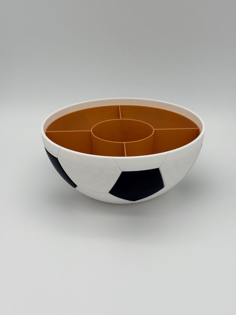May include: A round, bowl-shaped snack organizer designed with a soccer ball pattern on the exterior, featuring white and black pentagonal panels. The interior is divided into five distinct sections with a central circular compartment in brown.