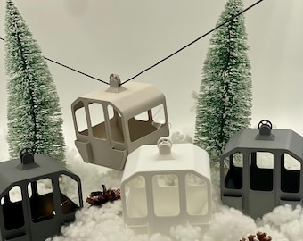 Puede incluir: Cuatro adornos en miniatura de teleféricos en blanco mate, gris claro y gris oscuro, suspendidos de un cable negro. Las cabinas están dispuestas entre dos pequeños pinos verdes nevados, piñas dispersas y nieve artificial blanca.