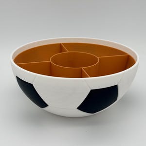 May include: A round serving bowl designed with a soccer ball pattern on the exterior, featuring white and black pentagonal shapes. The interior is a solid tan color and is divided into five separate compartments by a central circular divider.