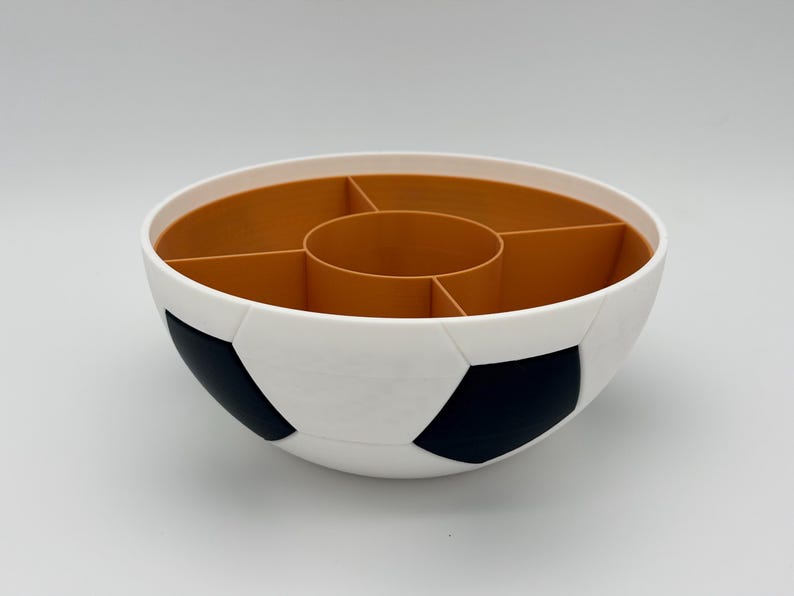 May include: A round serving bowl designed with a soccer ball pattern on the exterior, featuring white and black pentagonal shapes. The interior is a solid tan color and is divided into five separate compartments by a central circular divider.