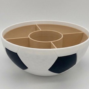 May include: A round bowl featuring a white and black soccer ball pattern on the exterior. The interior is light tan and divided into four outer compartments surrounding a central circular section, creating a segmented storage or serving container.
