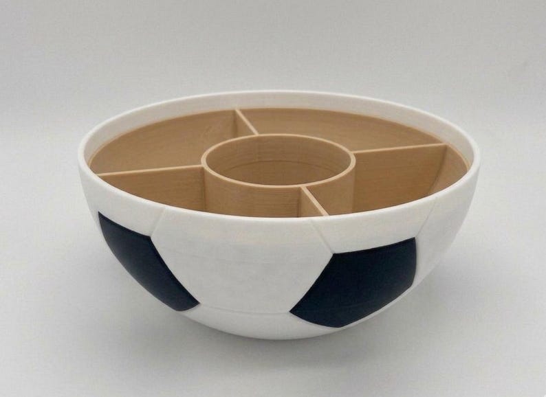 May include: A round bowl featuring a white and black soccer ball pattern on the exterior. The interior is light tan and divided into four outer compartments surrounding a central circular section, creating a segmented storage or serving container.