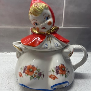 以下が含まれることがあります： 赤いフードをかぶった女の子の置物蓋が付いた、ビンテージのセラミック製のティーポット。ティーポットはオレンジ、パープル、グリーンの色合いの花柄で飾られています。ティーポットは底に青と白のトリムが付いています。