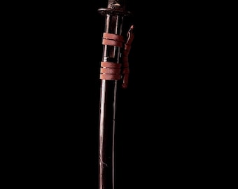 May include: A Japanese katana sword with a black handle wrapped in a black cord and a brown leather grip. The blade is dark with a slight curve. The sword has a brown leather tie near the handle.