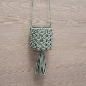 Könnte beinhalten: Ein hellgrüner Makramee-Pflanzenhänger mit Quaste. Der Hänger ist aus Baumwollschnur gefertigt und hat ein schlichtes Design.