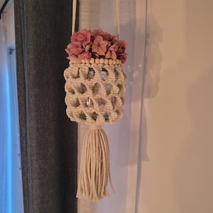 Könnte beinhalten: Ein Makramee-Pflanzenhänger mit einem weißen Seil und einem Holzperlenbesatz. Der Hänger hält ein Glasgefäß mit rosa Blumen.