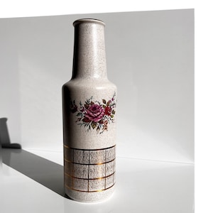 Puede incluir: Un jarrón de cerámica blanco con una superficie texturizada y un patrón de cuadrícula dorada. El jarrón está decorado con un diseño floral con rosas rosas y hojas verdes.