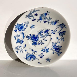 May include: A white porcelain saucer with a blue floral pattern. The saucer has a delicate design with small flowers and leaves.