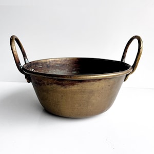 Könnte beinhalten: Eine kleine, antike Messingschale mit zwei gebogenen Griffen. Die Schale hat eine abgerundete Form und eine leicht angelaufene Oberfläche, die ihr ein Vintage-Aussehen verleiht. Die Griffe sind an den Seiten der Schale befestigt. Die Schale wird wahrscheinlich zum Servieren oder Ausstellen verwendet.