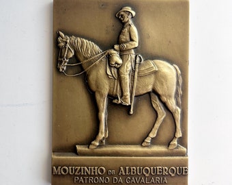 May include: A bronze plaque depicting a man on horseback. The plaque reads "Mouzinho de Albuquerque Patrono da Cavalaria".