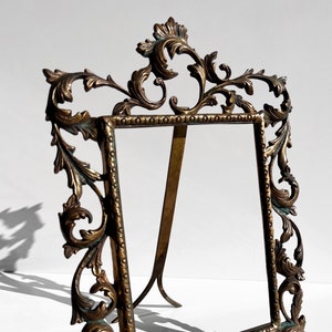 May include: A gold-toned metal picture frame with an ornate, scrolled design. The frame has a stand on the back and is designed to hold a vertical photo.