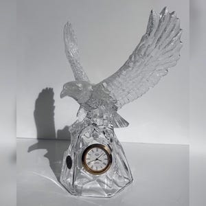 Puede incluir: Una figura de águila de cristal transparente con las alas extendidas, con un reloj con bisel dorado. La esfera del reloj muestra números romanos y manecillas. El águila está posada sobre una base facetada.
