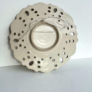 May include: White ceramic plate with a lace-like design and a hole in the center for hanging. The plate has a smaller, solid white plate in the center.