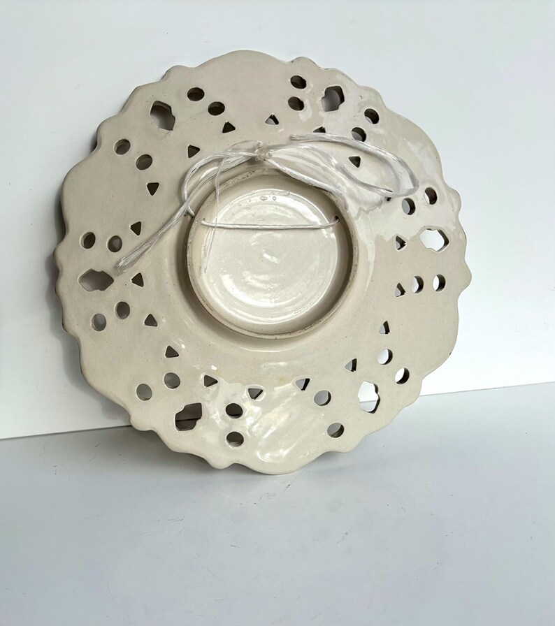 May include: White ceramic plate with a lace-like design and a hole in the center for hanging. The plate has a smaller, solid white plate in the center.