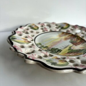 May include: A white ceramic plate with a scalloped edge and a lace-like design. The plate features a hand-painted scene of a house with a fence and trees. The plate has a hole pattern around the edge.