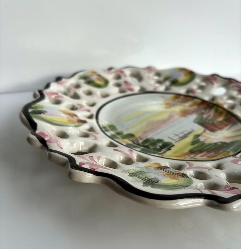 May include: A white ceramic plate with a scalloped edge and a lace-like design. The plate features a hand-painted scene of a house with a fence and trees. The plate has a hole pattern around the edge.