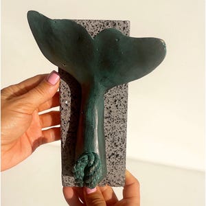 May include: A teal-colored whale tail sculpture mounted on a rectangular, textured gray base. The tail is curved and detailed, with a rope-like design at the base. The sculpture is held by hands with pink nail polish.