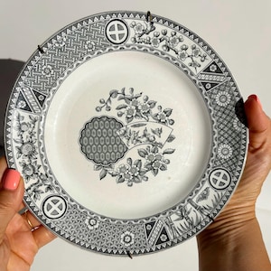 Puede incluir: Un plato de cerámica blanco con un patrón floral y geométrico negro. El plato presenta un diseño floral central y un borde decorativo con patrones y símbolos repetidos. El plato está sostenido por dos manos.