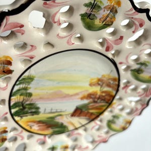 May include: A white ceramic plate with a hand-painted landscape scene. The plate has a scalloped edge and a delicate openwork design. The landscape features trees, a road, and a body of water. The plate is decorated with pink, green, and brown accents.