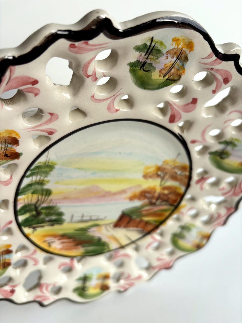 May include: A white ceramic plate with a hand-painted landscape scene. The plate has a scalloped edge and a delicate openwork design. The landscape features trees, a road, and a body of water. The plate is decorated with pink, green, and brown accents.