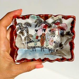 May include: Rectangular ceramic dish with a red, scalloped border and a gold trim. The dish features a painted scene of figures on a bridge, with a house and trees in the background. The colors include red, white, black, and gold.