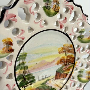 May include: A white ceramic plate with a lace-like design around the edge. The center of the plate features a hand-painted landscape scene with trees, mountains, and a lake. The plate is decorated with pink, green, and yellow accents.