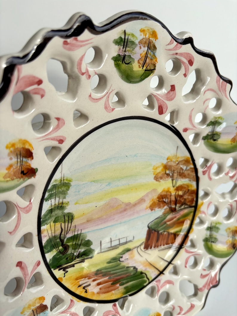May include: A white ceramic plate with a lace-like design around the edge. The center of the plate features a hand-painted landscape scene with trees, mountains, and a lake. The plate is decorated with pink, green, and yellow accents.