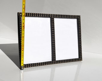 Könnte beinhalten: Ein rechteckiger, schwarz-goldener Doppelbilderrahmen mit zwei weißen Flächen für Fotos. Der Rahmen ist etwa 40 cm hoch. Der Rahmenrand hat ein strukturiertes, genietetes Design.