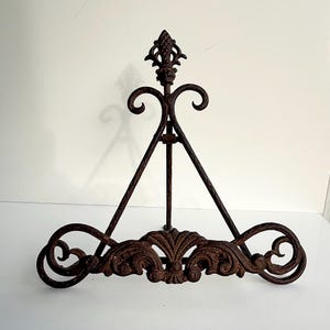 Peut inclure: Chevalet décoratif en fer forgé avec une finition marron vieillie. Le chevalet présente des volutes ornées et un fleuron en forme de fleur de lys. Le cadre triangulaire supporte une étagère inférieure avec des fioritures décoratives.