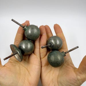 Könnte beinhalten: Vier antike Metallknöpfe mit einer verwitterten, grünlich-grauen Oberfläche. Jeder Knopf ist kugelförmig, mit einem Gewindestab, der sich zur Befestigung nach hinten erstreckt. Ein Knopf hat eine flache, kreisförmige Basis. Die Knöpfe werden in offenen Händen gehalten.