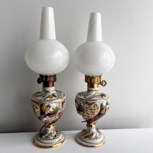 Op de afbeelding: Twee witte porseleinen olielampen met gouden accenten en bloemmotieven. De lampen hebben witte glazen kapjes en staan op een wit oppervlak.