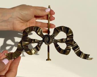 May include: A vintage brass bow-shaped decorative object. The bow has detailed ribbon folds and a central oval embellishment. It is held by a vertical rod, suggesting it may be a decorative handle or accent piece. The brass has a warm, aged patina.