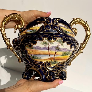 May include: A decorative, navy blue ceramic container with gold-colored handles and accents. The container features a landscape painting with trees, a river, and mountains. The handles are ornate and curved.