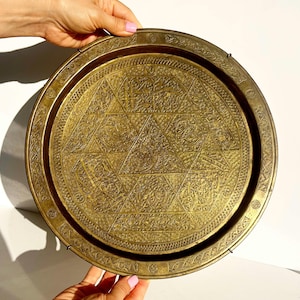 May include: A round, antique brass tray with intricate geometric and floral patterns. The tray features a raised edge and a central design composed of interlocking triangles. The brass has a warm, aged patina, suggesting its vintage origin.