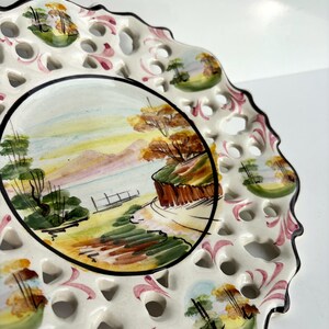 May include: White ceramic plate with a hand-painted landscape scene of trees, hills, and a body of water. The plate has a scalloped edge and a delicate lace-like design around the rim.